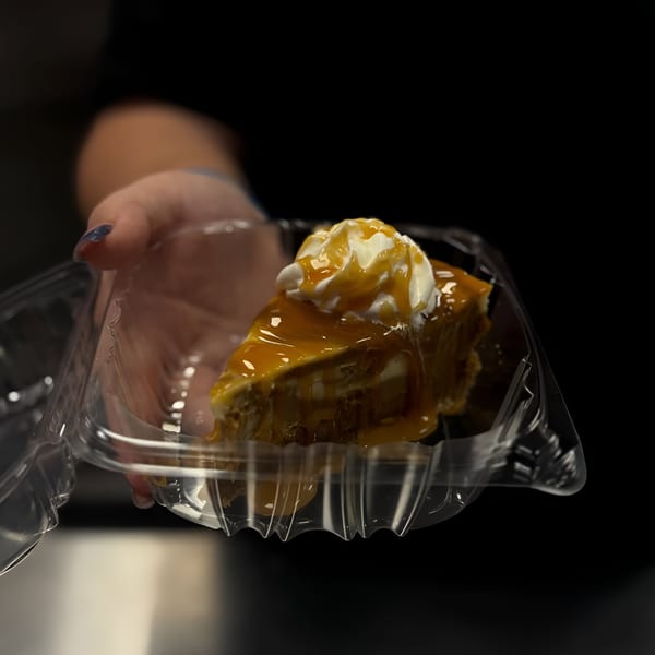 A hand holds a clear plastic container with a slice of cheesecake topped with whipped cream and caramel.