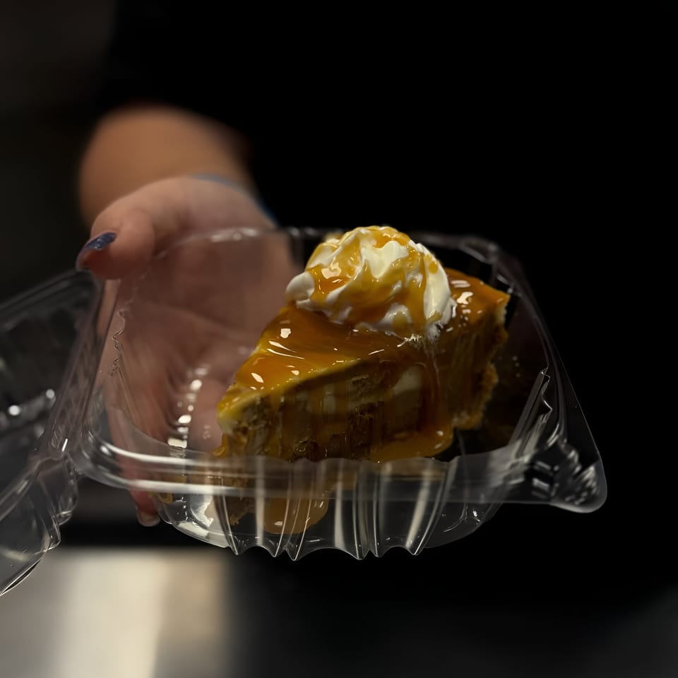 A hand holds a clear plastic container with a slice of cheesecake topped with whipped cream and caramel.