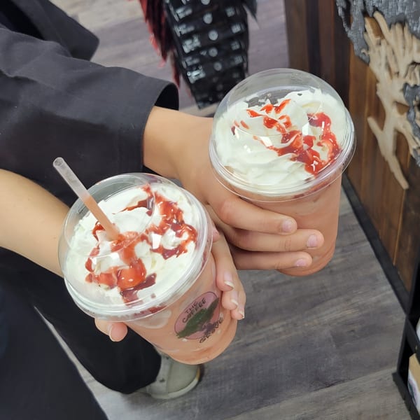 Two hands holding The Coffee Grove Corp strawberry frappes with whipped cream and syrup.