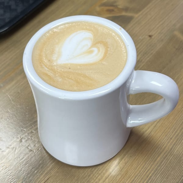 A white mug of latte with a heart-shaped foam design sits on a wooden table.